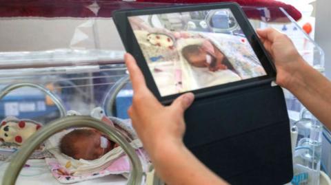 A woman holding up an ipad to a baby incubator, with a baby inside, and she appears to be on on a video call 