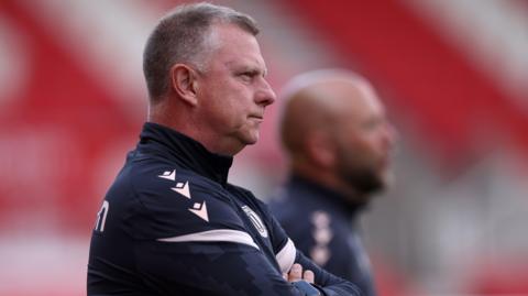 Stoke boss Mark Robins stands with arms folded looking out from the touchline during a match