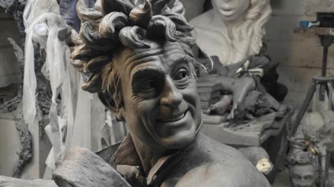 A close up picture of the clay model used for the sculpture showing Terry Jones with wild hair and smiling. He wears only a tie that looks like it is blowing in the wind