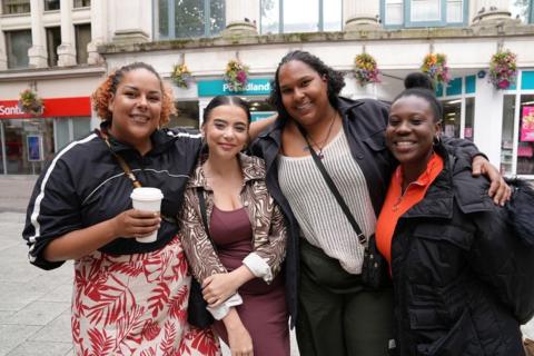 Four women, standing with their arms round each other in front of a Poundland shop. One is holding a hot drinks cup.