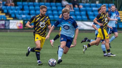 A footballer in a blue kit and football boots chases a football on a green pitch. A player in a yellow and black strip follows. Players from both other teams can also be seen in the background.