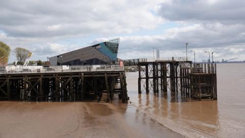 A side-view of Victoria Pier. The water is murky and the sky is overcast with white clouds. The structure is made up of large brown wooden beams and in the background is a large aquarium.