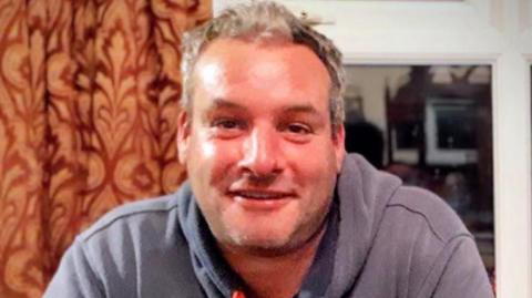 A picture of a man with a grey hoodie and who has black and grey hair. He is sat down. He is sat in front of an orange curtain. 