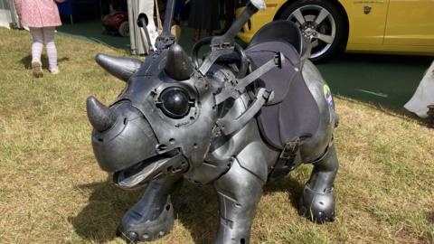 A robotic looking dinosaur stands in a grass field. It is a dark grey colour and has three horns on its head. It has a black saddle fitted to its body.