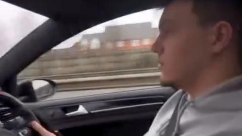 A man wearing a grey hooded top is sat in a car, holding a steering wheel, as he is focused on the road