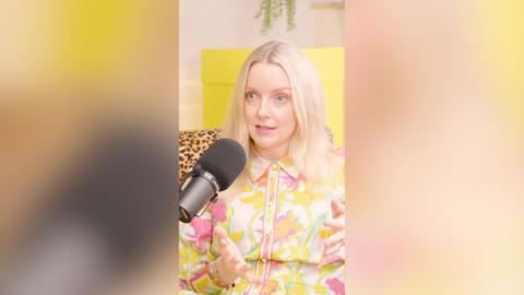 A woman with shoulder-length blonde hair wearing a floral shirt sits in front of a microphone and talks to someone off-camer