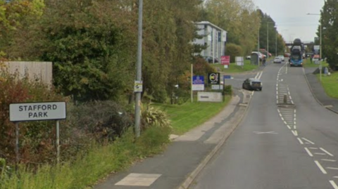 A white road sign that reads "Stafford Park" on the side of a road. The road is has two single carriageways and goes off into the distance. There is a junction of the left which a car is turning into. A bus, which is driving towards you, is approaching in the distance.
