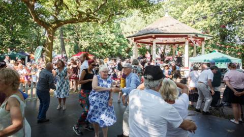 Crowds of people at the festival in 2024. In the foreground is a dancefloor area with couples jive dancing. In the background is a band playing in a bandstand.