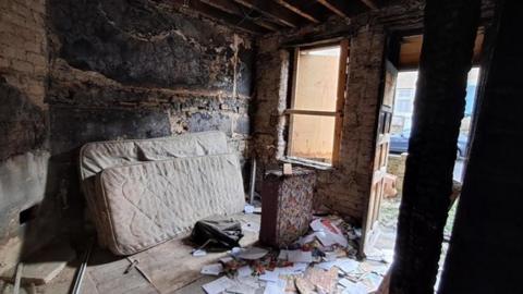 The interior of a severely damaged and abandoned room. The walls are blackened and charred. The ceiling beams are exposed, and the wooden structure appears burned and deteriorated. There is a dirty, stained mattress leaning against the wall, and the floor is littered with scattered papers, debris, and what looks like a piece of clothing or fabric. A small, damaged piece of furniture stands near the centre of the room. The window and door openings are boarded up with wooden planks, but some daylight is coming through. Outside the door, part of a street and a parked car are visible.