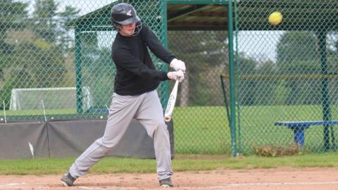Joseph Partridge is on the pitch with a bat, wearing a black helmet. A yellow softball can be seen in motion as it flies off the bat