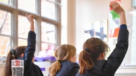 Three teenage girls sitting in a classroom with their backs to the camera. Two of them are raising their hands to answer a question. There are pencil cases, folders and a copy of Romeo and Juliet sitting on the desk in the foreground.