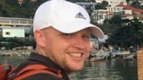 A smiling man wearing a white cap with a black adidas logo on it and a black polo shirt. Behind him is some water and houses.