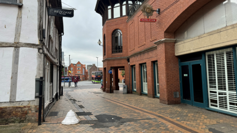 A pedestrianised section of St Stephens Church Lane running between an old, timber-framed building and a modern, brick-built shopping centre