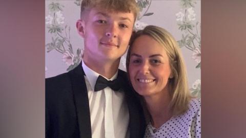 A teenage boy with short blond hair is wearing a black and white suit with a black bow tie and is standing next to a woman with shoulder length blonde hair with a white top with black polkadots on it. They are both smiling against a flower wallpaper backdrop.