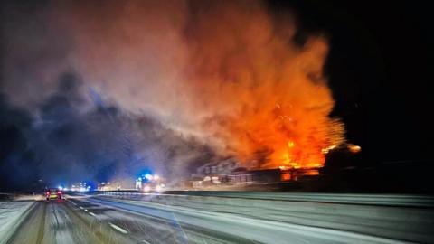A snowy road with a large fire engulfing houses close to the carriageway with orange flames and smoke