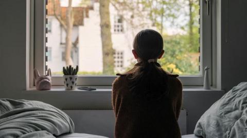 The back of a young girl sat on a bed staring out of a window.