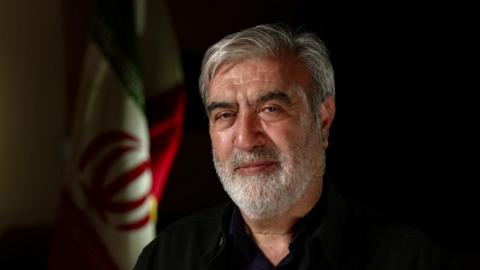 A man with a grey beard and hair looks into the camera. He is wearing a black shirt and behind him is the Iranian flag.
