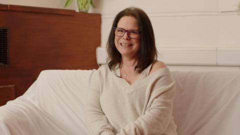Paula Van Santen smiling for an image in a room. She is sitting down on a cream couch and is wearing a cream top.