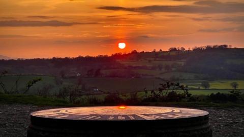 The sun is setting as a white ball in orange and yellow skies over hill and fields in the distance and a huge sundial in the foreground