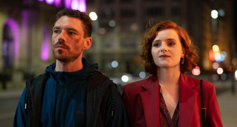 Filming of This City is Ours with Michael Kavanagh, played by James Nelson-Joyce, with short brown hair and beard, and Diana Williams, played by Hannah Onslow, with shoulder length red hair wearing a red blazer and burgundy top. There are walking in the street in the evening holding hands.