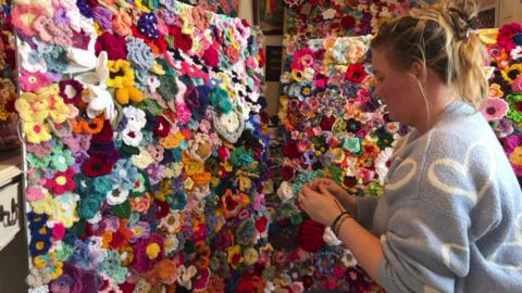 Thousands of brightly coloured crochet flowers being pinned to a large pin boards. A woman is in the forefront working on attaching the flowers to the board. 