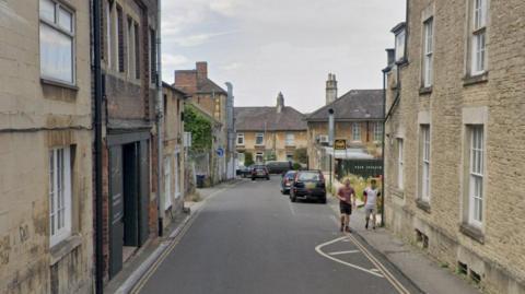 A view of Union Road in Chippenham on an overcast day. A single-lane road is surrounded by cream-coloured houses. Several cars are parked on the side of the road and two people are walking on the right-hand side.