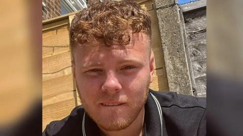 A young man with short strawberry blonde hair and a nose piercing wearing a black polo shirt