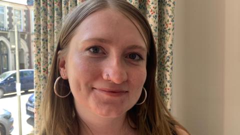Marianne looks at the camera, she's wearing hoop earrings and has a nose piercing. She's smiling and a curtain and window overlooking the street is visible behind her.