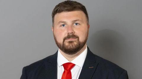 Head and shoulders picture of Blackpool South Labour MP Chris Webb. He has short dark hair and a short beard and is wearing a navy suit, white shirt and red tie