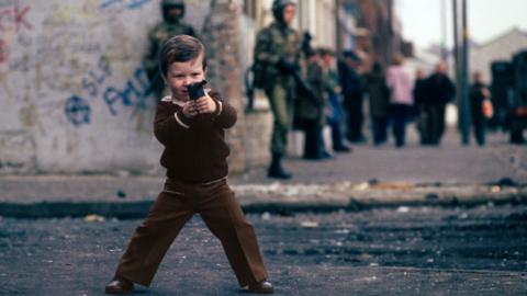 A small boy in 1970s clothes points a toy gun at the camera. There is graffiti on a wall behind him.