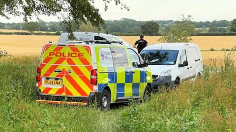 Three police vans parked on a field, with an officer looking away into the field.