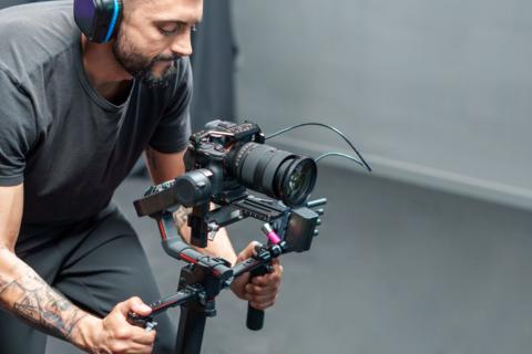 A man with tattoos on his arms is wearing headphones. He is dressed in a grey T-shirt and grey trousers and is operating a small movie camera on a rig.