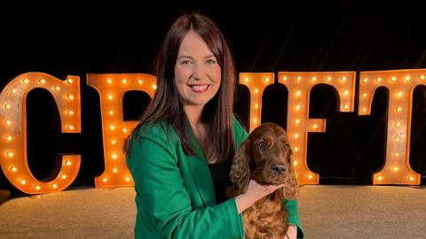 Laura Crombie sitting in front of a lit up Crufts sign kneeling on the floor with her Irish setter called Evie. She has a green blazer on with long straight brown hair. She is smiling into the camera 
