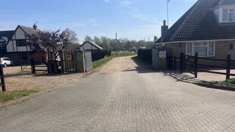 Two silver gates are in between two houses. Both of them are open and a driveway leads to a field. There is a 10mph sign on a pillar on the right and a defibrillator is on the left gate.