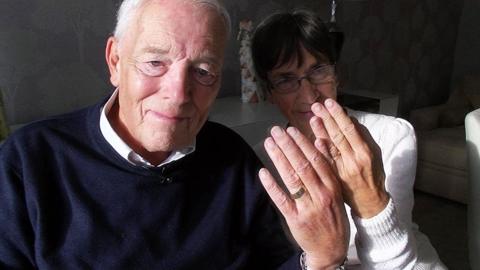 Christopher Brightmore has receding grey hair and blue eyes. He is wearing a blue jumper and is sitting next to his wife, who is wearing a white jumper and metal, oval-shaped spectacles on her face. They are both holding their hands up to the camera showing their wedding rings, which they are wearing on their ring finger.