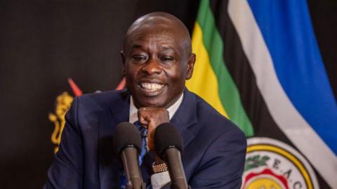 Deputy President of Kenya, Rigathi Gachagua speaks into microphones at a press conference at his official residence in Nairobi, on October 7, 2024. He is wearing a navy suit with a flag behind him.