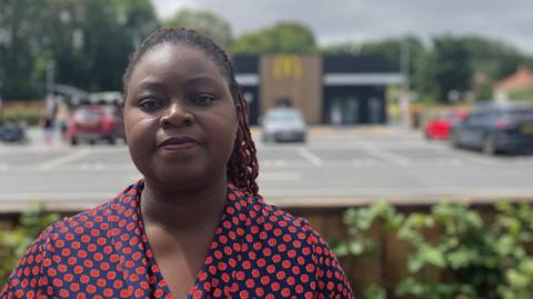 Makala Zulu looks towards the camera wearing a red and blue patterned top, with a McDonald's restaurant blurred in the background