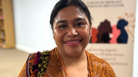 A woman with tied back black hair smiles into the camera. She wearing a traditional East Timorese yellow dress and a orange, black, pink and black shawl over her shoulder. She is wearing an orange beaded necklace.