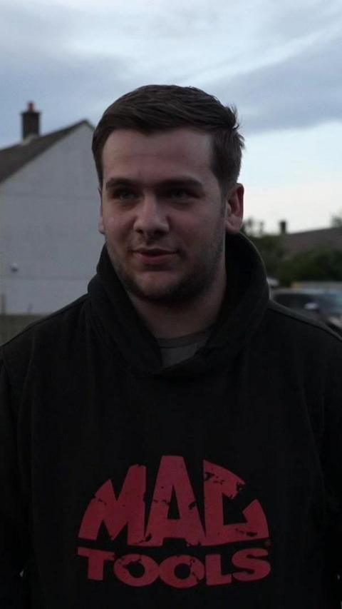 The image shows a man with brown hair. He is wearing a black hoodie. Behind him are cars and houses.