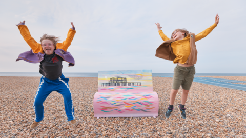 Two children jumping in the air on Brighton's shingle beach in front of one of the book bench sculptures which depicts the city's burnt out west pier. The bench is pink with multi coloured waves
