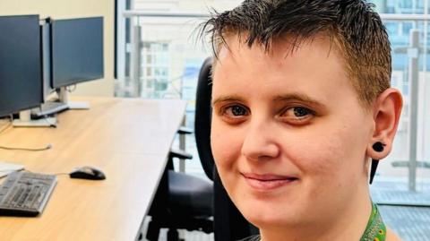 Ashleigh Bishop smiles at the camera. She has short hair and sits in an office environment, with computer screens behind her. It is a portrait shot, and lanyards at the top of her neck can be seen, including a sunflower one for invisible disabilities