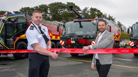 Two men stood in front of three fire emergency response vehicles, holding up and cutting a red ribbon. One man is holding up the ribbon while the other is using scissors to cut it. They are both smiling and looking straight at the camera.