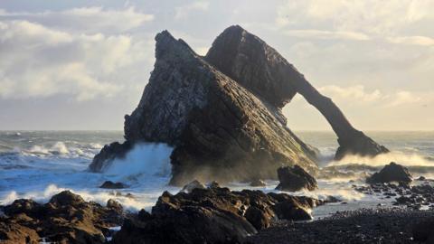 A striated rock formation coming out of a dramatic sea right at the shore. It is sunny.