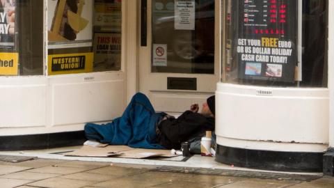 A man in a sleeping bag in shop door front in York. He is smoking and lying on flattened cardboard boxes.