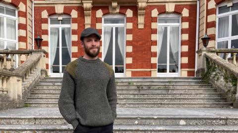 A man wearing a grey jumper and black trousers, stood on a set of concrete steps. Behind him is a red-bricked building with tall windows with draped curtains.