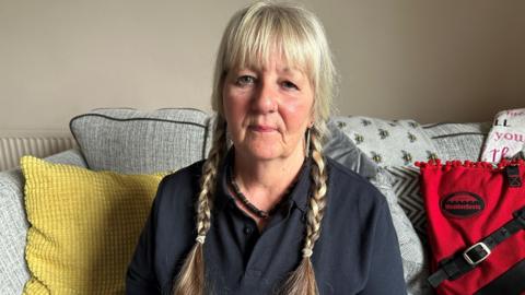 Susan O Sullivan, with blonde plaits in her hair and wearing a black top, is sitting on a grey sofa with a yellow cushion.