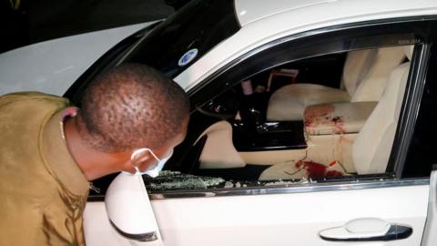 A detective wearing a mask examines a white car with broken windows and some blood on the passenger seat  