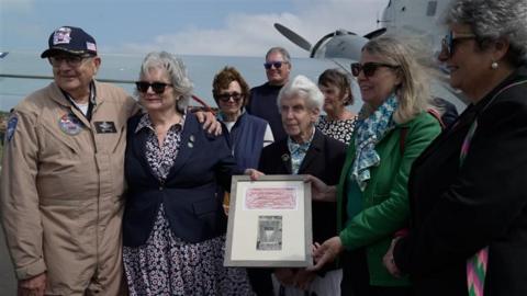 Group photo with a man in a pilot uniform on the left. Two ladies with grey hair and navy blazers hold on to a frame containing a red crayon rubbing and photo.
