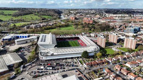 A drone photo of Ashton Gate, taken from a very high angle. It shows crowds going into the stadium, which is in the middle of a built-up area. The River Avon and Clifton Suspension Bridge are visible in the background. 