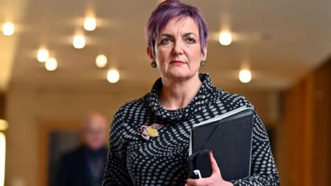 A woman with short hair dyed purple is wearing a black and white spotted top. She is holding a tablet and phone in her left hand as she was through a corridor